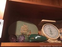 Tin can with a sailing ship painted on, American Family dial scale, two purple glass door knobs, and green ocarina seen on wooden shelf.
