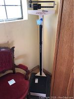 Full view of the Detecto mechanical scale showing the large black base platform, central vertical white post with height ruler, and balance bar with sliding weights at the top.