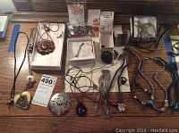 Photo showing full assortment of costume jewelry laid out on wooden table including necklaces, pendants, small boxes and loose jewelry pieces.