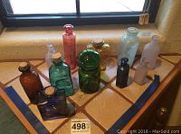 Photo showing 11 apothecary style glass bottles in assorted colors on a tiled floor near a window.