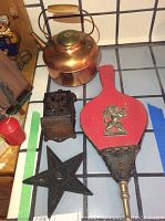 Four vintage items on white tiled surface: copper tea kettle, cast iron match holder, red and black bellows, and black metal star.