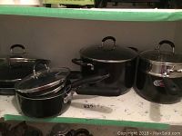 Photo showing multiple black pots and pans with glass lids arranged on a shelf, displaying various sizes and black handles.