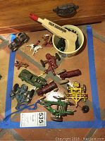 Photo shows a group of vintage cast iron toys including toy cars, figures on horses, and a toy gun labeled 'Colt Director'. Various colors including green, blue, red, cream, and black are visible. Toys show wear and paint chipping.