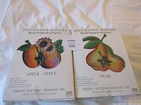 Front view of both boxed Decorative Hanger units showing apple and pear artwork and product details