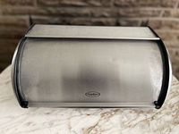 Front view of stainless steel bread box showing roll-top door and logo badge