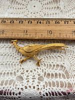 front view of gold-tone pheasant brooch beside ruler showing length