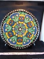 Front view of hand-painted glazed ceramic plate on display stand with measuring tape showing approx 11 inch diameter