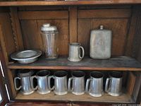 Cabinet shelves holding pewter tankards, bowl, glass shaker, flask