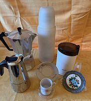 Group shot of all items including two moka pots, thermos, grinder, one cup & saucer, and one coaster