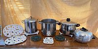 Full lot showing three stainless stockpots, roaster and assorted trivets