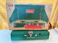 Closed green Coleman 2-burner stove sitting below original printed cardboard box