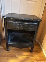 Front view of black wood nightstand showing drawer, lower shelf, scalloped apron, cabriole legs, worn paint