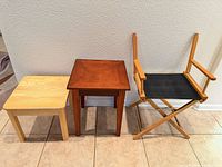 Three furniture items aligned against wall: light wood table, brown wood table, director chair frame