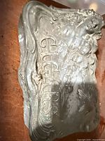 Top view of decorative metal cigar box with raised "CIGARS" lettering, female figure and floral motifs, showing dent and patina