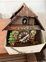 Front view of chalet cuckoo clock inside box showing dial, cuckoo door, painted trim, chains