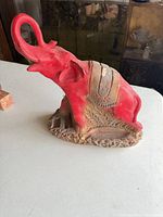 Front left view of red plaster elephant with raised trunk showing decorative saddle and base