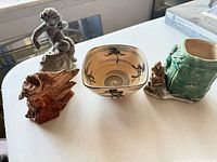 All four items grouped on table: metal cherub figurine, carved wood piece, beige ceramic bowl, green planter with small figurine
