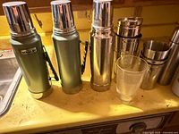 Two Stanley green thermoses, stainless thermos, stacking cups and tumbler on counter