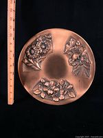 Front of round copper plaque with floral relief and ruler showing size