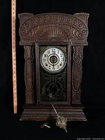 Front view of carved wooden mantel clock with ruler for scale, pendulum and key visible