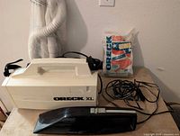 White Oreck XL canister vacuum, black Oreck XL 12 V vacuum, filter bags and hose on table