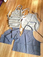 Stack of beige aprons, one beige apron opened, several gray aprons opened, showing ties and pockets