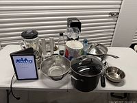 Overall table view of kitchen items including blender, SodaStream, cookware, colander, stock pot, skillet and paper plates