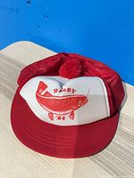 Front view of red and white Darby '84 trucker hat showing fish graphic and pom-pom