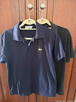 Front view of navy and black Lacoste polo shirts on hangers