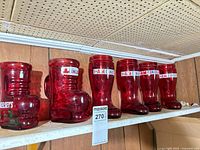 Six red glass boot beer glasses lined on shelf showing logos and shape