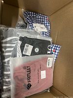 Top layer of box showing pink Famolly sweatshirt, black top, blue gingham shirt all sealed