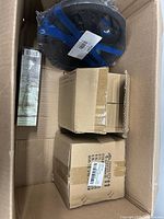 Open carton showing black circular item, tall slender boxed item and sealed brown boxes