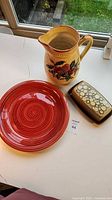 Overall view of red plate, fruit pitcher, butter dish on white surface