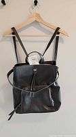 Front view of faux leather backpack purse on hanger showing flap, front zip pocket, straps