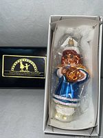 Sealed ornament in open front cardboard box next to black lid with gold logo