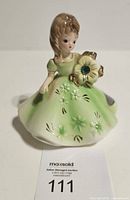 Front view of May figurine showing dress, flower, rhinestone