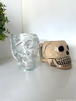 Clear glass and beige ceramic skull steins side by side, profile view