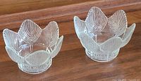 Two clear pressed-glass lotus votive holders on wooden surface