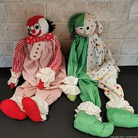Both cloth clown dolls side by side against brick backdrop