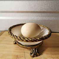 Stone egg resting inside footed milk glass bowl on metal stand