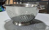 Side profile of stainless steel colander showing perforations, loop handle and footed base