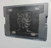 Top view of black laptop cooling pad showing perforated metal surface, central fan and rubber grips