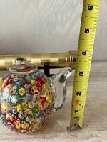 Glass teapot paperweight beside tape showing height, brass kaleidoscope in background