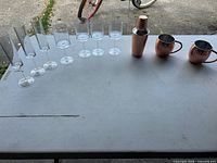 All drinkware and barware items laid out: 5 plastic champagne flutes, 6 plastic wine glasses, 1 copper shaker, 2 copper mugs