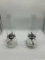 Front view of two matching clear glass Lamplight 110 oil lamps showing bases, burners, wicks and chimneys