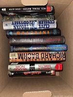 Top view of nine hardcover novels inside a cardboard box, spines visible showing titles and authors