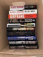 Stack of 10 hardcover fiction books inside cardboard box showing spines and titles