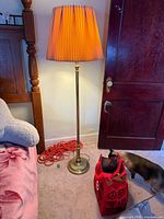 Full view of brass floor lamp standing lit beside bed, showing base, column, and pleated shade