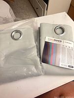 Two folded ChrisDowa curtain panels, one in clear plastic, showing grommets and label
