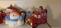 Row of ceramic Christmas teapots and sugar bowl, showing snowman and pickup-truck forms